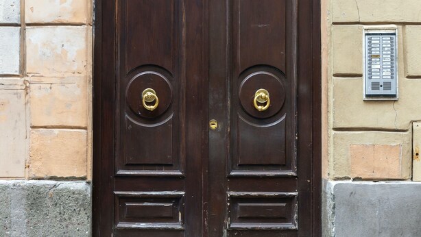 Une porte à deux battants en bois foncé, ornée d’une poignée de porte en laiton, est encadrée dans la façade d’un immeuble aux couleurs beige et gris.