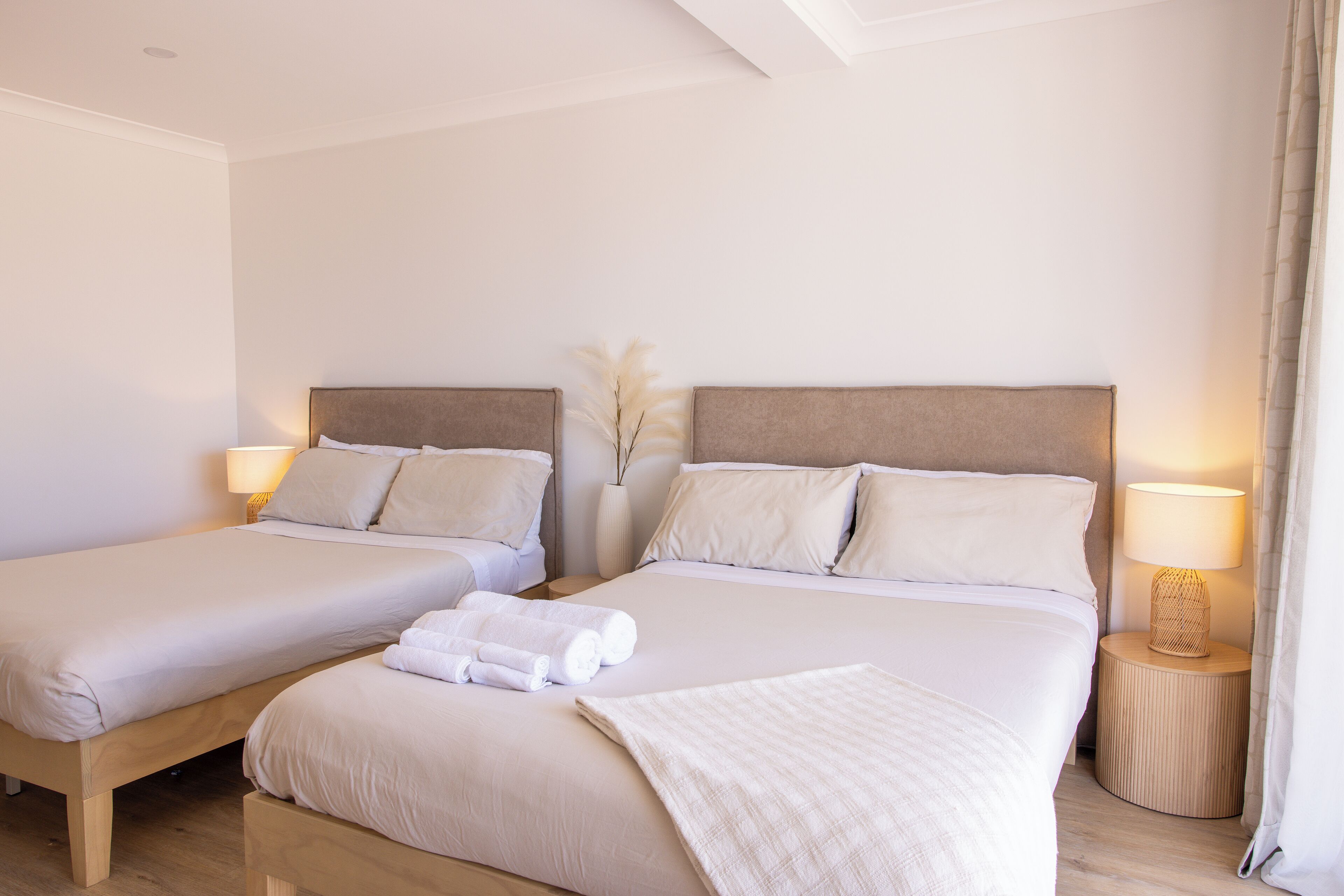 A hotel room with two beds, each with a headboard, bedside tables with lamps, and a vase with dried flowers on the wall.