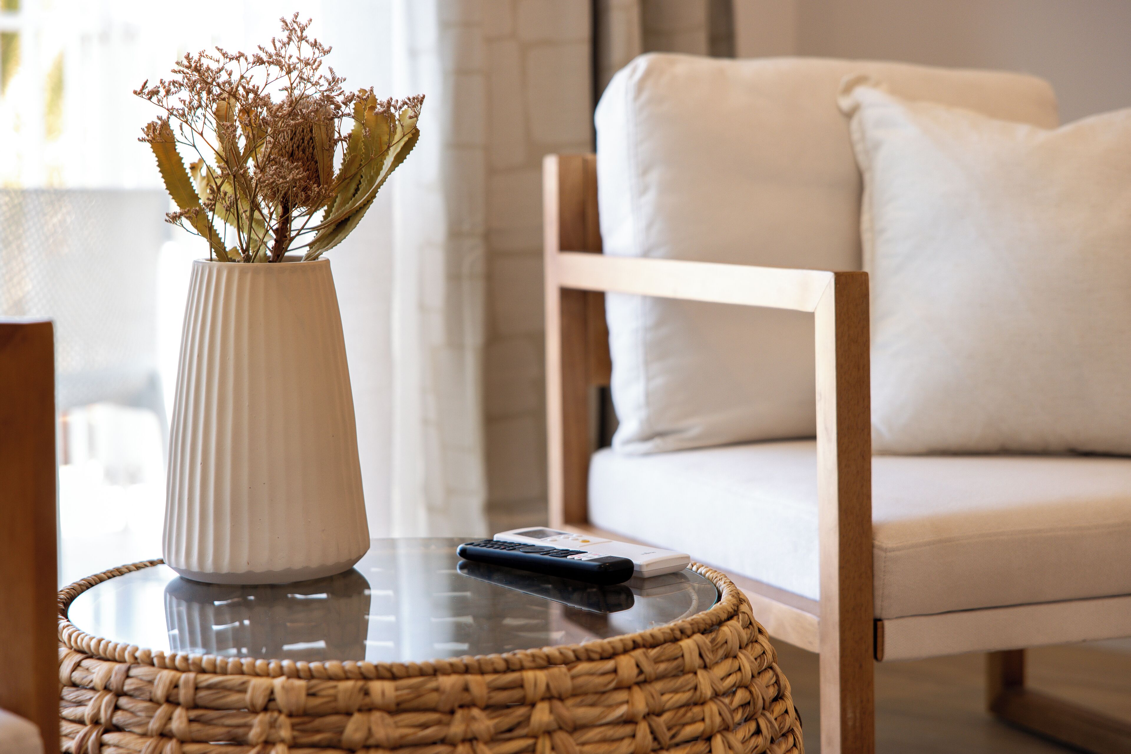 A white vase with dried flowers on a glass table, a remote control, and a wicker basket.