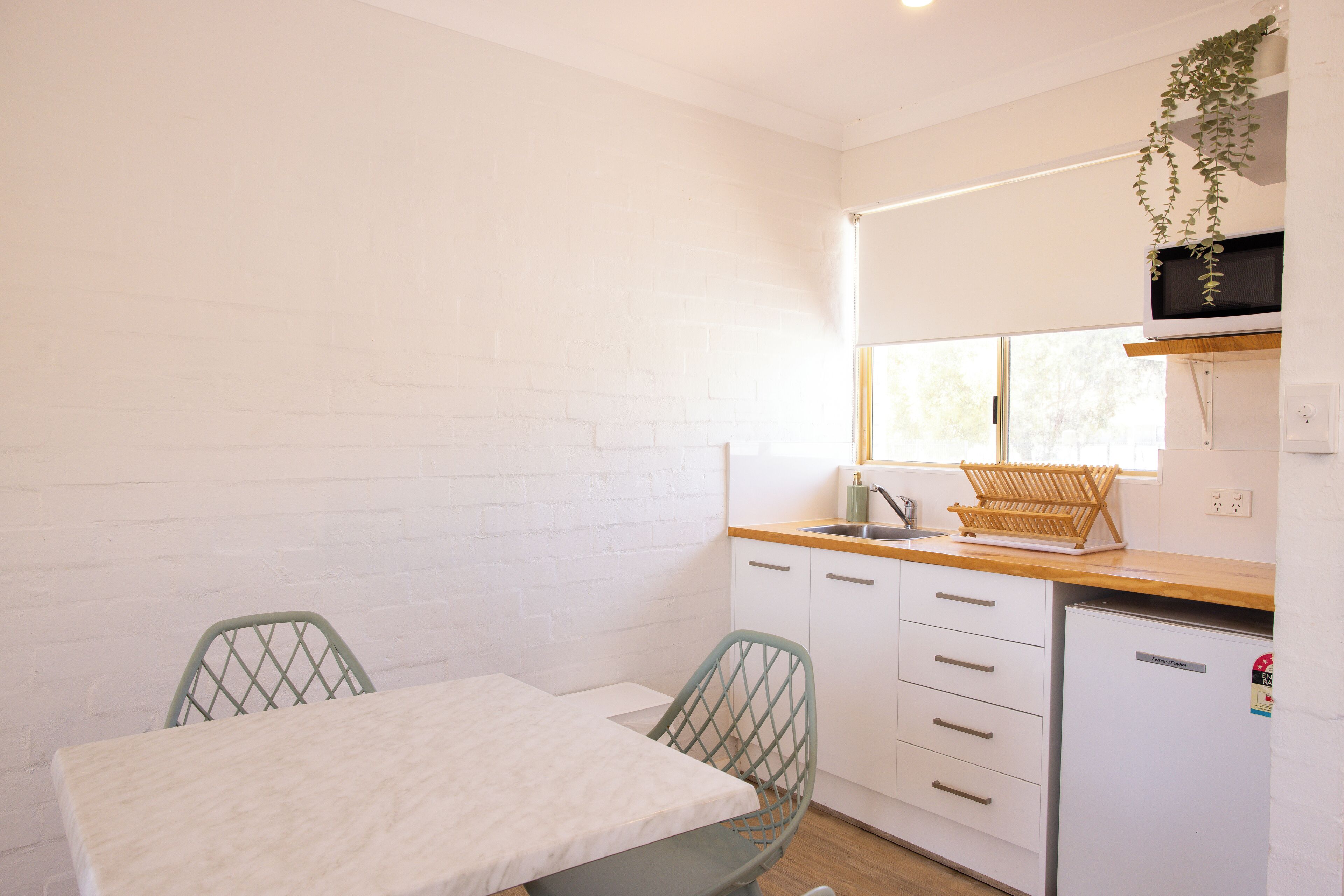 A compact kitchen with white brick walls, a wooden countertop, a small refrigerator, and a window with a plant hanging above the sink.