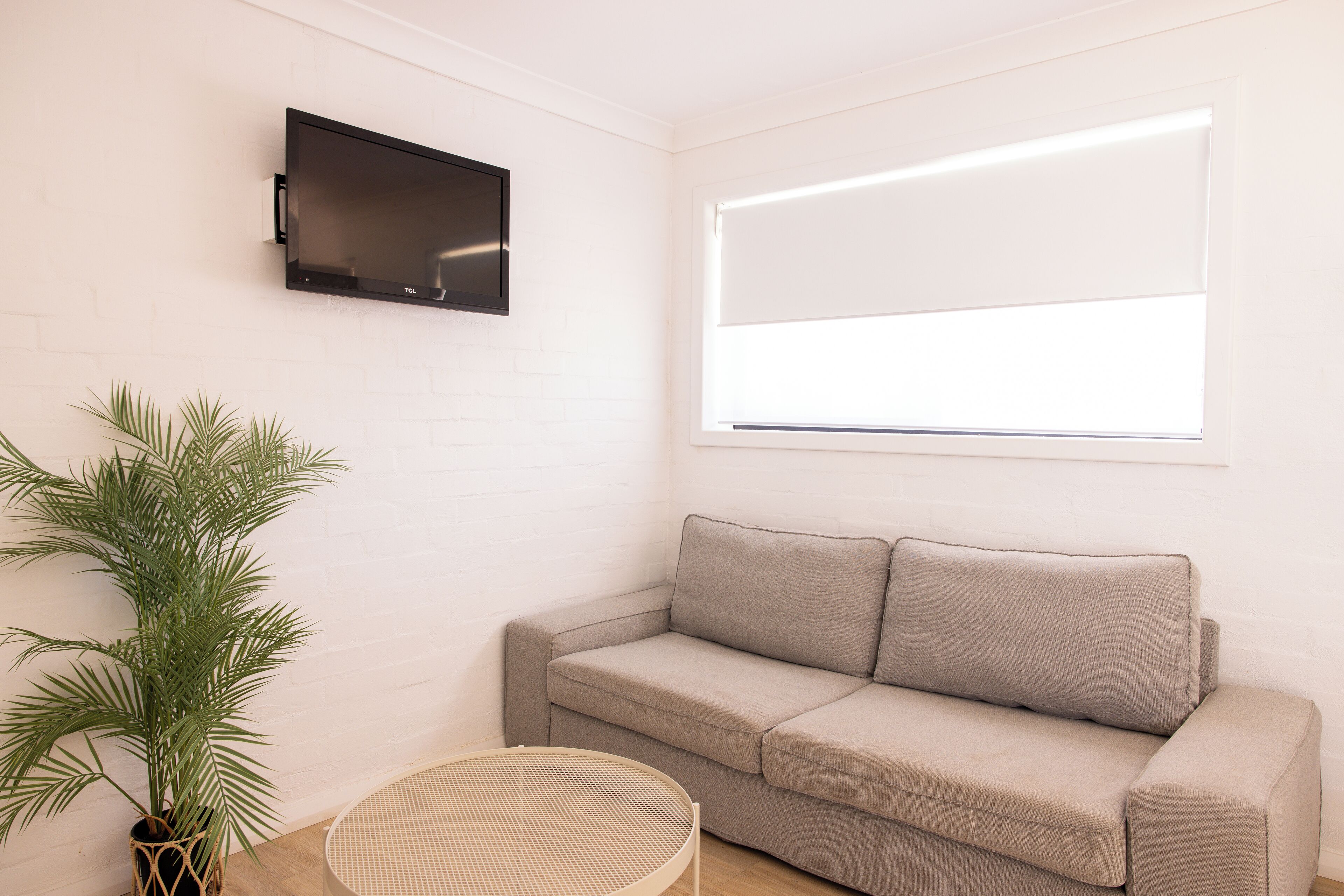 A room with a beige sofa, a flat-screen TV mounted on the wall, a potted plant, and a wicker chair.