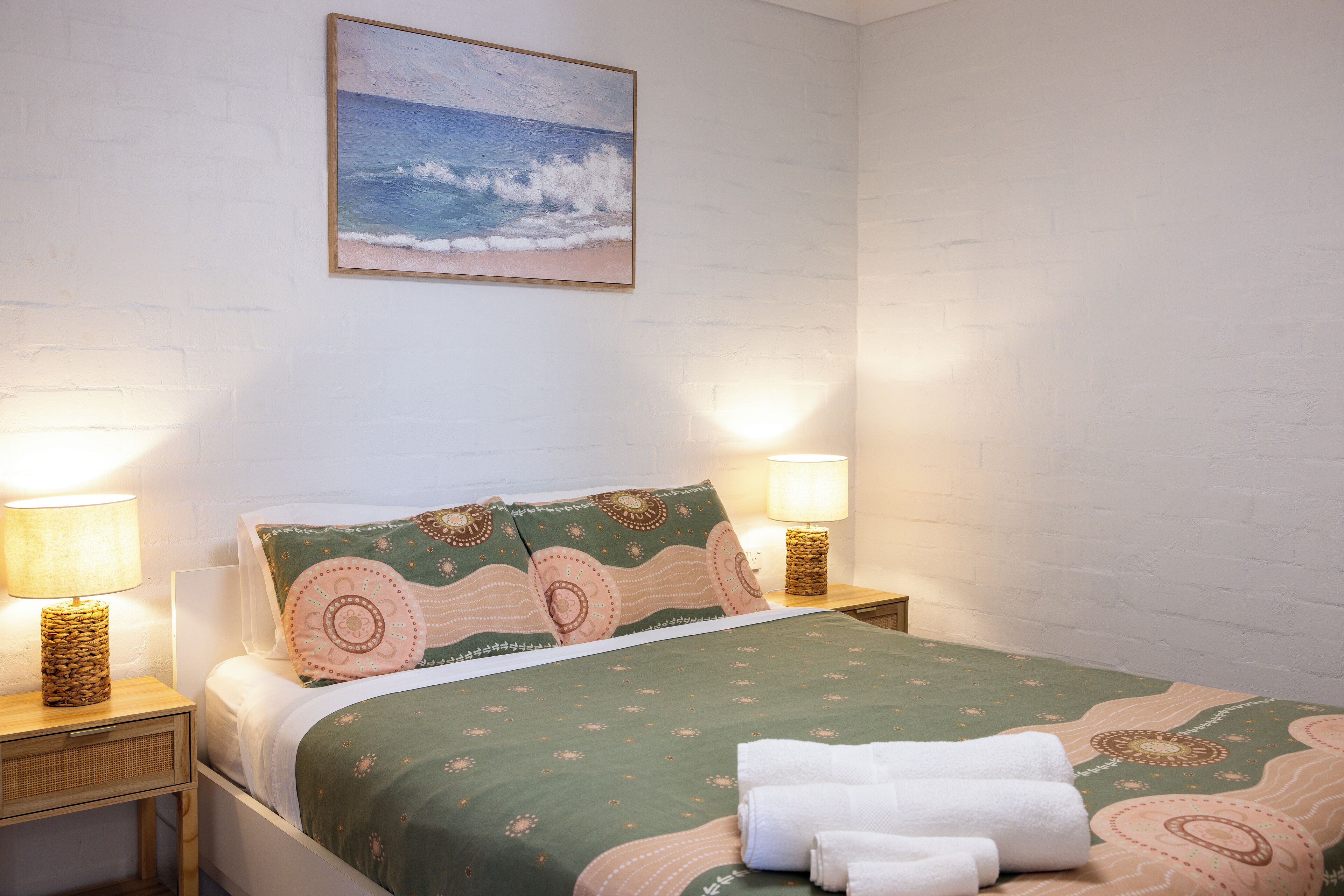 A neatly made bed with a patterned bedspread, two bedside lamps, and a painting of the ocean on the wall.