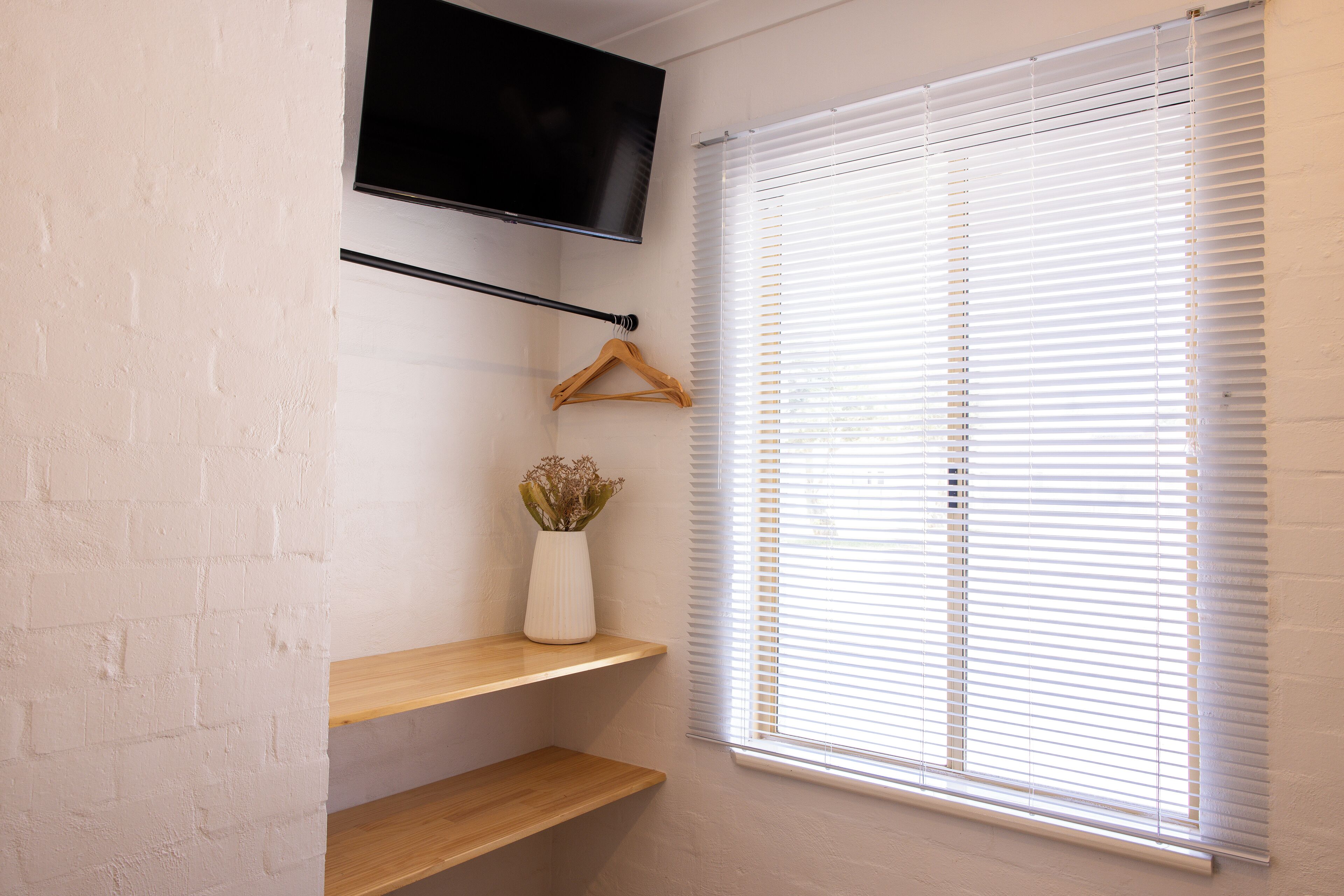 Une pièce avec un téléviseur fixé au mur, une étagère en bois, un porte-manteau suspendu et une fenêtre avec des stores.