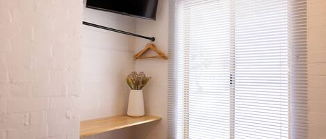 A room with a TV mounted on the wall, a wooden shelf, a hanging coat rack, and a window with blinds.
