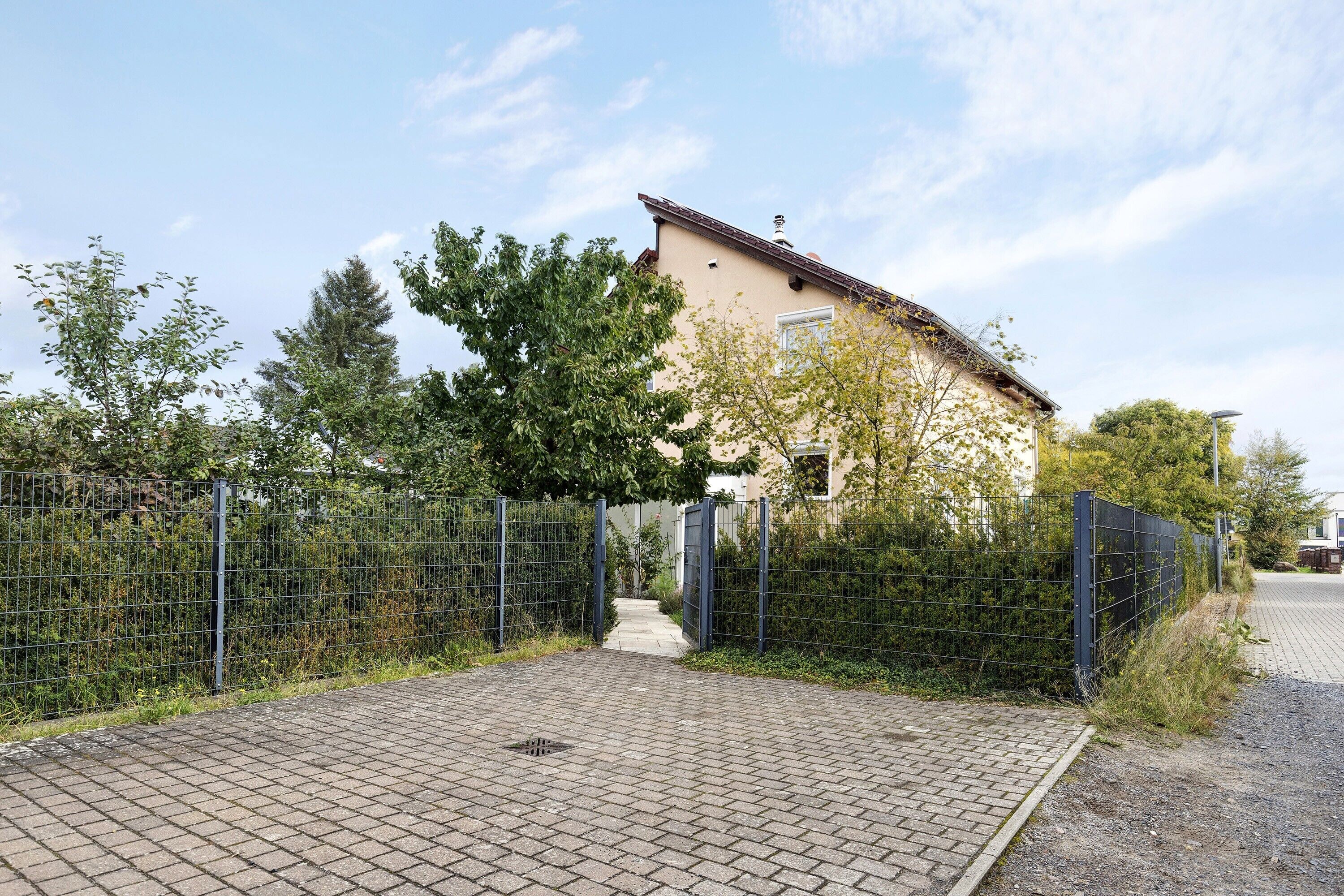 A paved driveway leading to a house with a metal gate, surrounded by greenery and a clear sky.