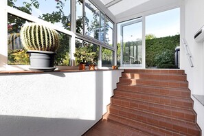 Eine Treppe führt zu einem Wintergarten mit großen Fenstern, einem Topf mit einem Kaktus und Blick ins Grüne.