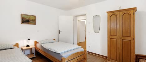 A bedroom with two single beds, a wooden wardrobe, a desk with a lamp, and a painting on the wall.
