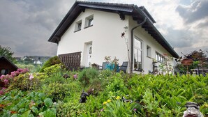 Una casa blanca con techo inclinado, un porche de madera y un jardín con diversas plantas y flores.