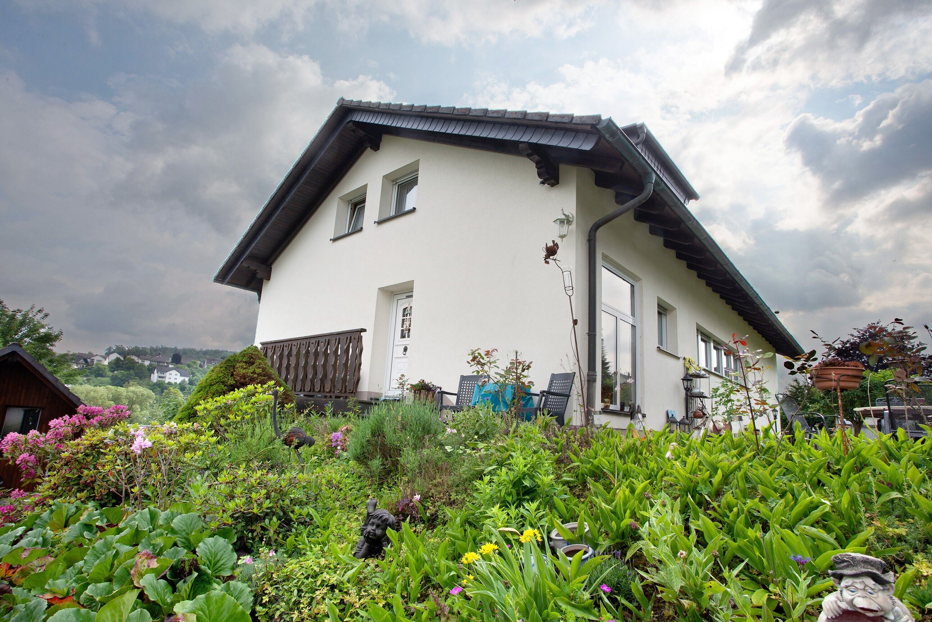 Une maison blanche avec un toit en pente, une terrasse en bois et un jardin où poussent différentes plantes et fleurs.