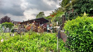 Un edificio rústico de madera con un porche cubierto, rodeado de abundante vegetación y diversas decoraciones de jardín.