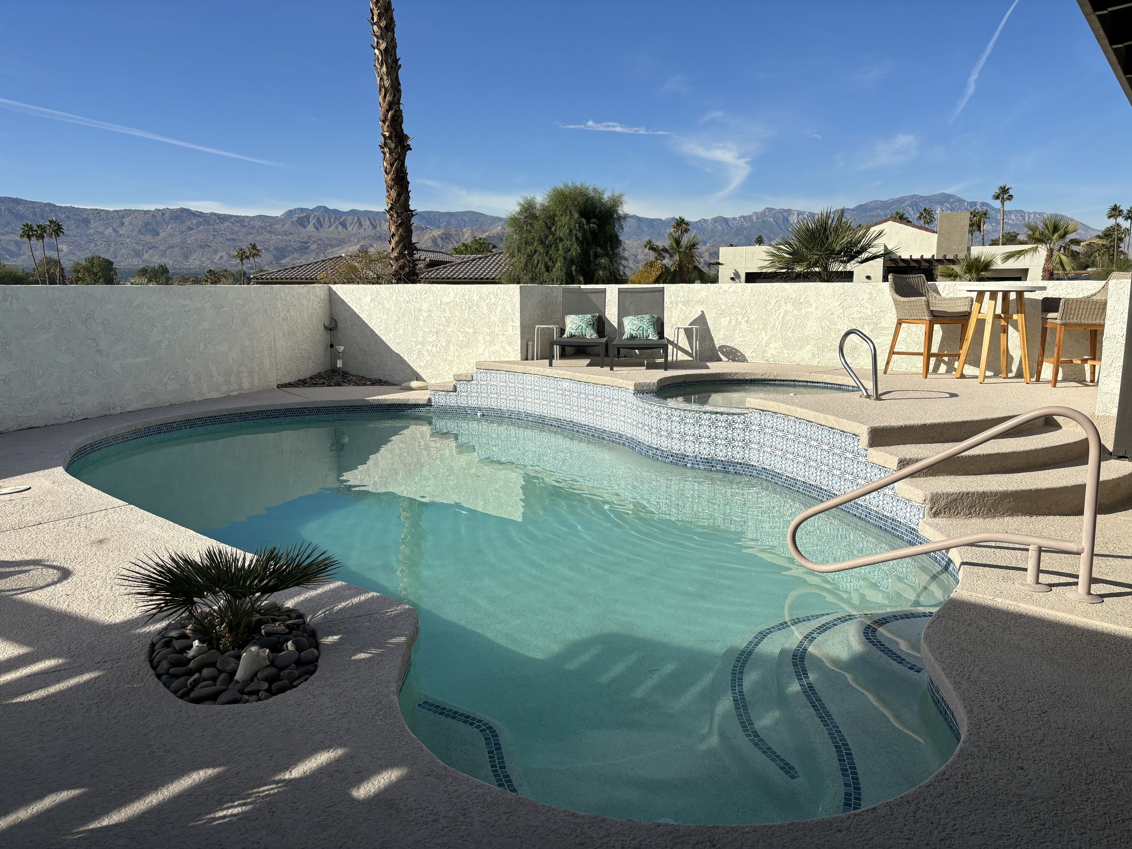 Una piscina con borde de azulejos, rodeada por un muro blanco y palmeras. Hay escalones que bajan a la piscina y un patio con sillas y una mesa.