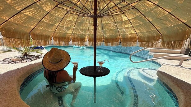 A person relaxing by a circular pool with a thatched umbrella, a small table with a drink, and a stone pathway leading to the pool.