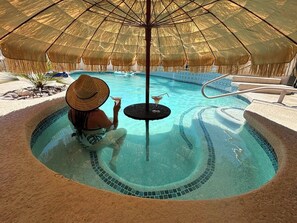 A person relaxing by a circular pool with a thatched umbrella, a small table with a drink, and a stone pathway leading to the pool. - Desert Oasis with Pool + Mtn Views! + Golf (Palm Desert)
