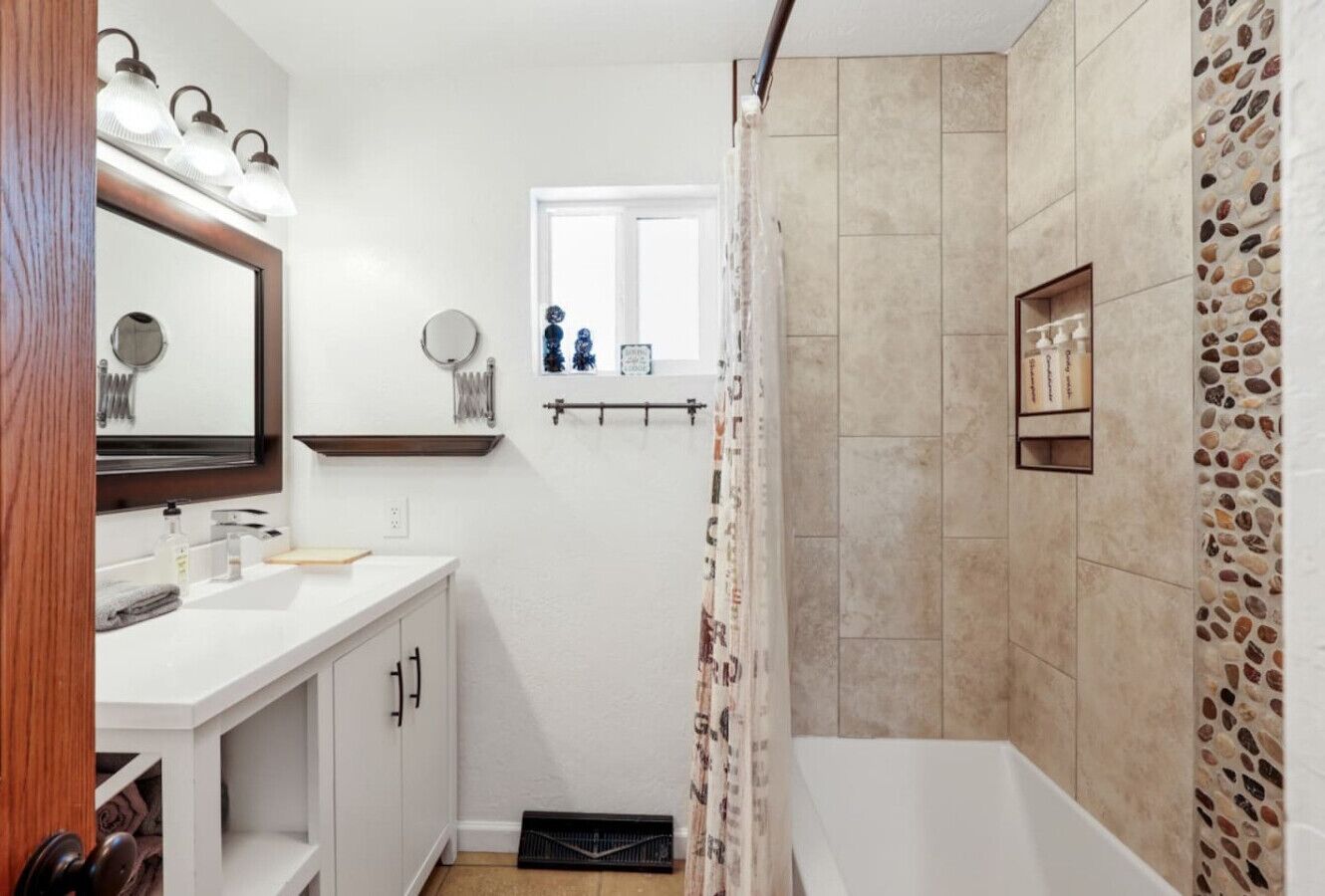 Een badkamer met een wit wastafelmeubel, spiegel en een douche met beige tegels, en een raam boven de wastafel.