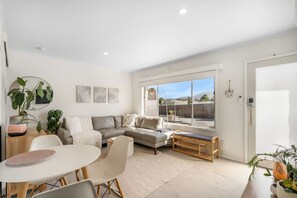 Una sala de estar moderna con un sofá seccional gris, una mesa de centro redonda y un comedor con una mesa y sillas blancas. Hay varias plantas, un ventanal con vistas, y una puerta con cerradura.