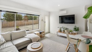 Een moderne woonkamer met een grote schuifdeur naar een achtertuin, een flatscreen televisie aan de muur en een eethoek met een kleine tafel en stoelen.