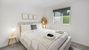 Een slaapkamer met een bed, nachtkastje, lamp en een raam met uitzicht op groen.