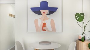 Een kamer met een witte muur, een lijst met een afbeelding van een vrouw met een blauwe hoed, een ronde witte tafel en twee witte stoelen.