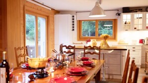 A wooden dining table set with red plates, wine glasses, and a bottle of wine. There are chairs around the table, and a window with a view of greenery is visible. - Chalet Le Haut Pré by Interhome (Villars)