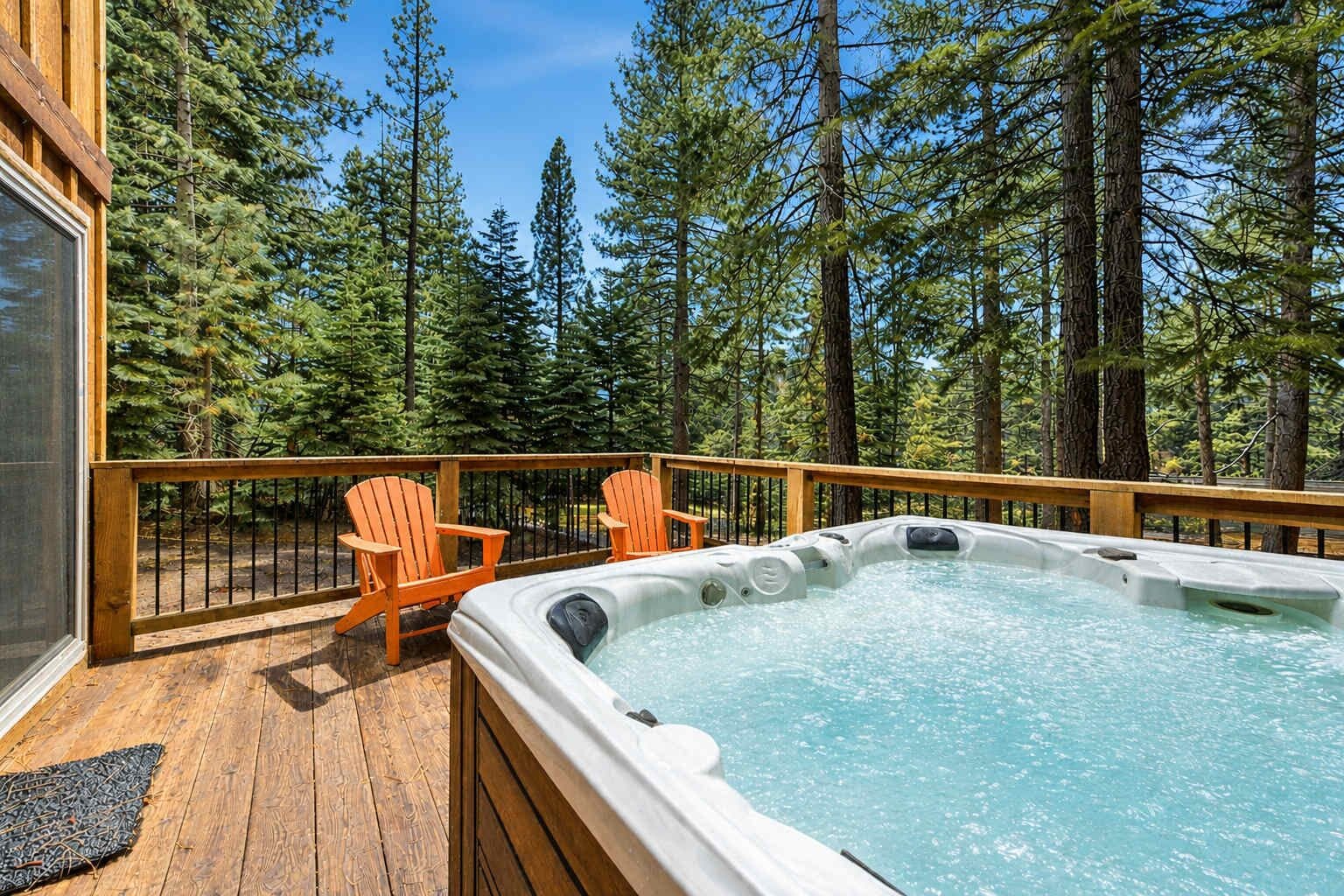 A hot tub on a wooden deck with two Adirondack chairs, surrounded by tall pine trees.