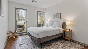 A bedroom with a bed, two windows, a bedside table with a lamp, and a patterned rug.