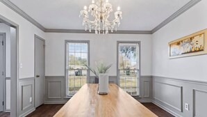 A spacious room with a long wooden table, a chandelier, and large windows with blinds.