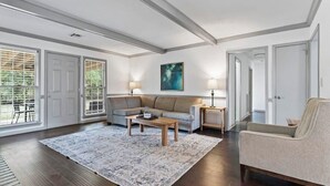 A spacious living room with a large sectional sofa, a coffee table, and a patterned rug. There are two lamps on side tables, and a painting on the wall.