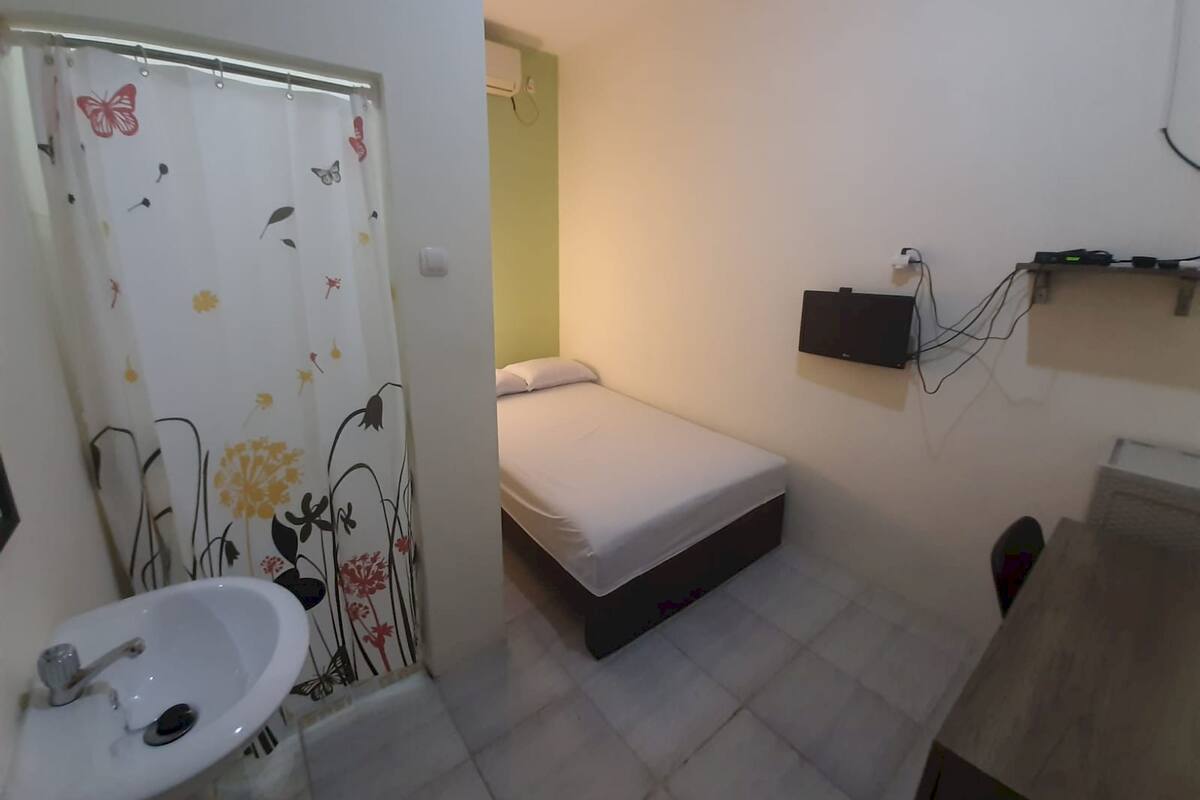 Ein kleines, kompaktes Zimmer mit einem Einzelbett, einem Nachttisch, einem an der Wand montierten Fernseher und einem Badezimmer mit Waschbecken und Duschvorhang.