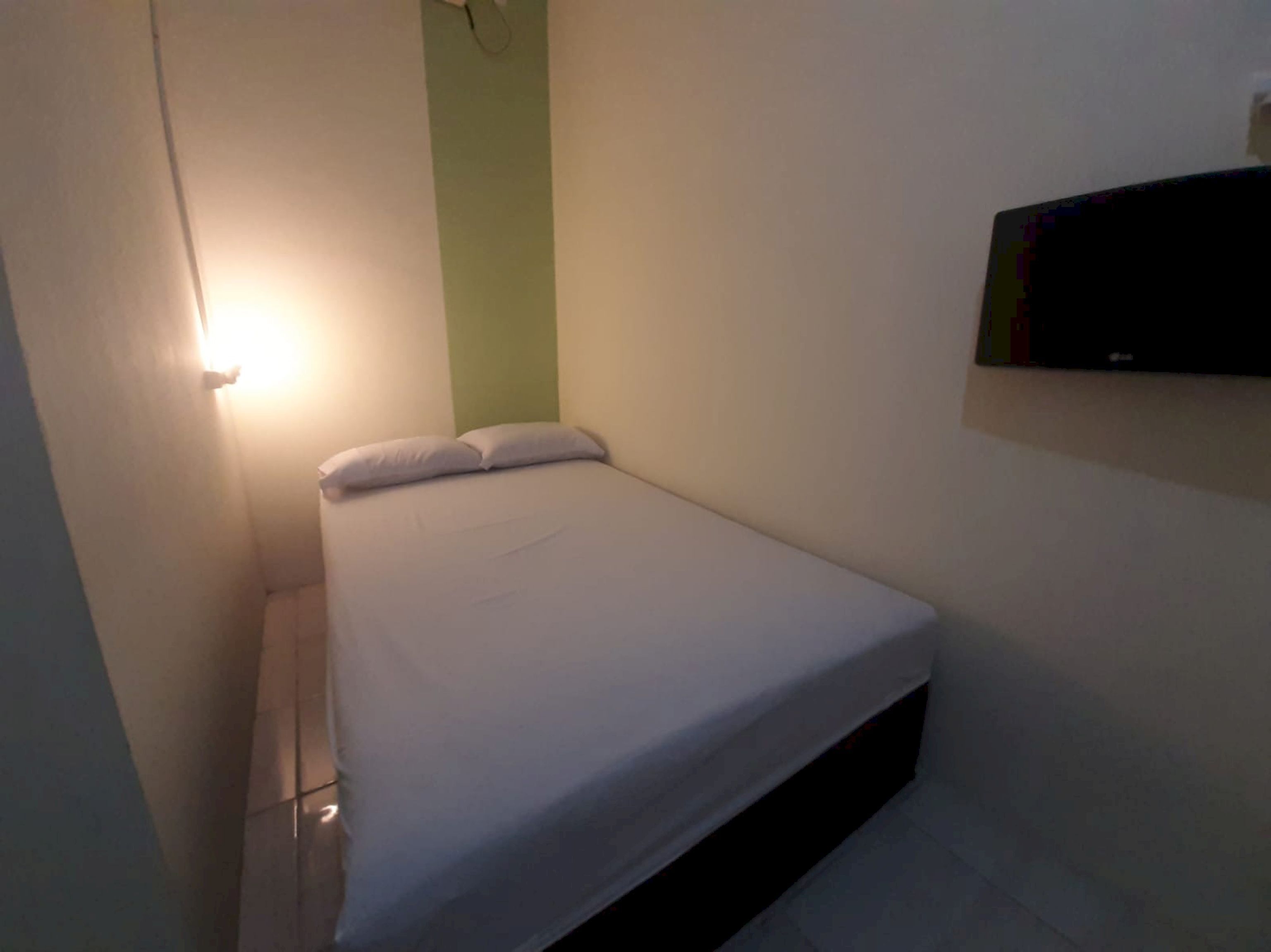 A small, dimly lit room with a single bed, a green wall, and a TV mounted on the wall.