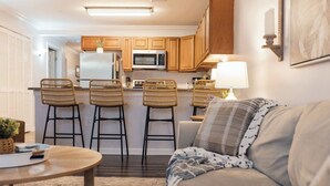 A compact living space with a kitchen area featuring wooden cabinets, a microwave, and a refrigerator. There are three wicker chairs and a sofa with patterned cushions. A table with a remote control and a plant is visible.