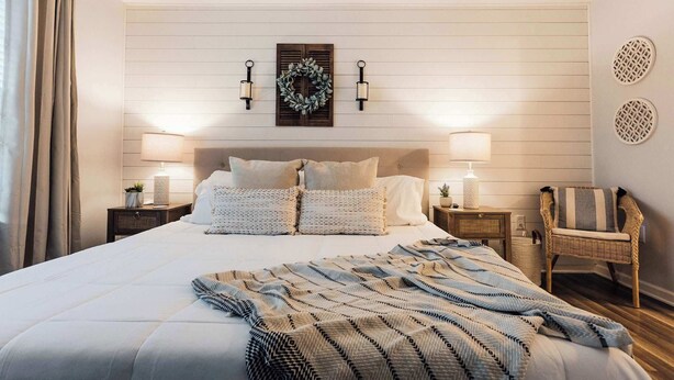 A neatly arranged bedroom with a large bed, two bedside tables with lamps, a decorative wreath on the wall, and a wicker chair.