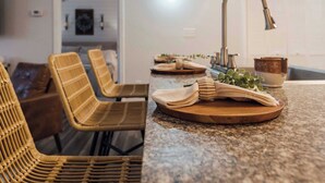 A kitchen with a granite countertop, wicker chairs, a wooden serving board with cloths, and a modern faucet.