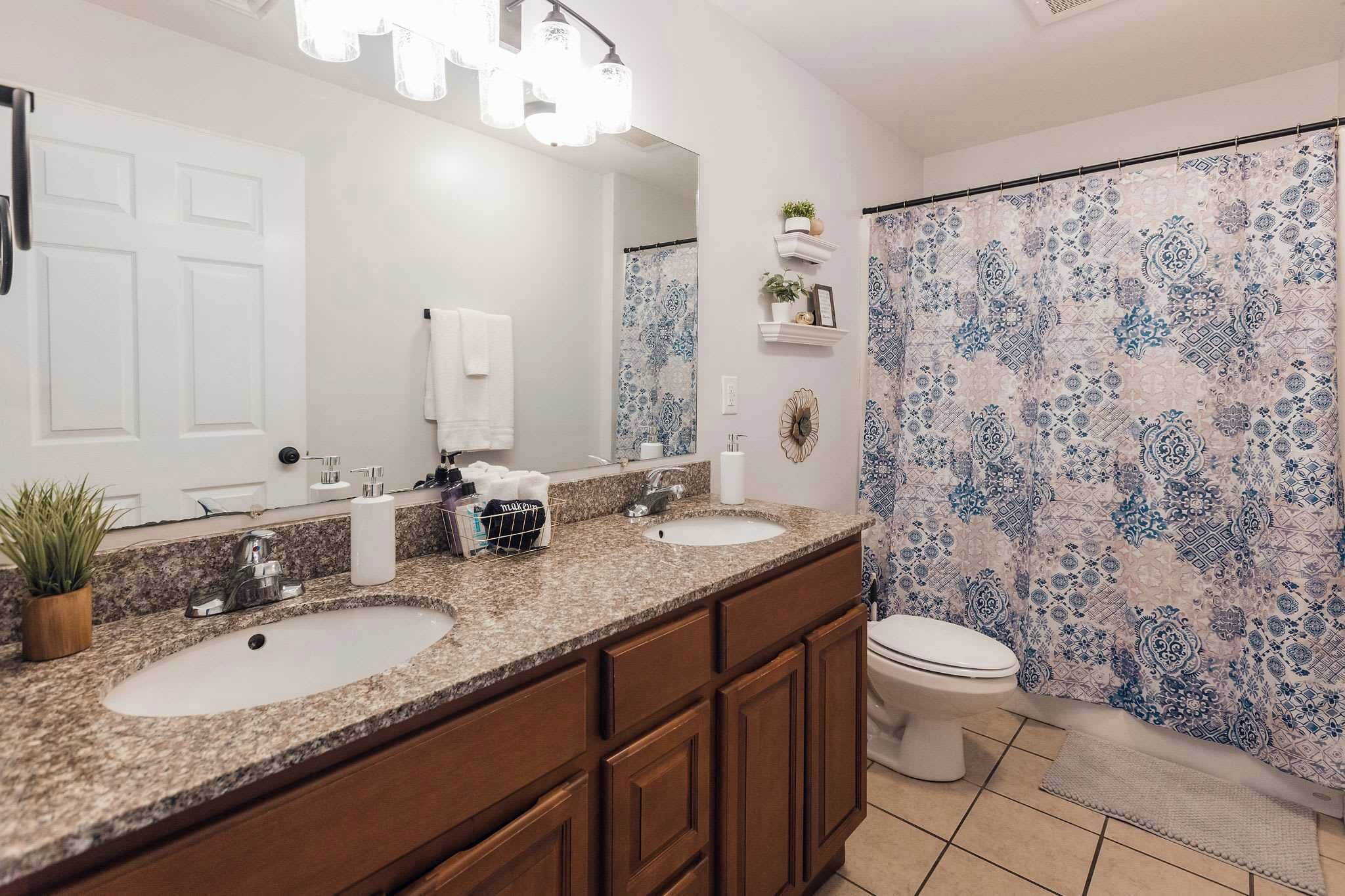 Une salle de bain avec deux lavabos, des toilettes, un grand miroir et un rideau de douche à motifs bleus et blancs.