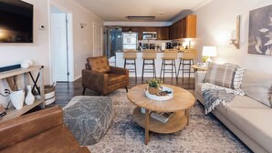 A living room with a sofa, armchair, coffee table, and a TV. There is a kitchen area with a dining table and chairs.