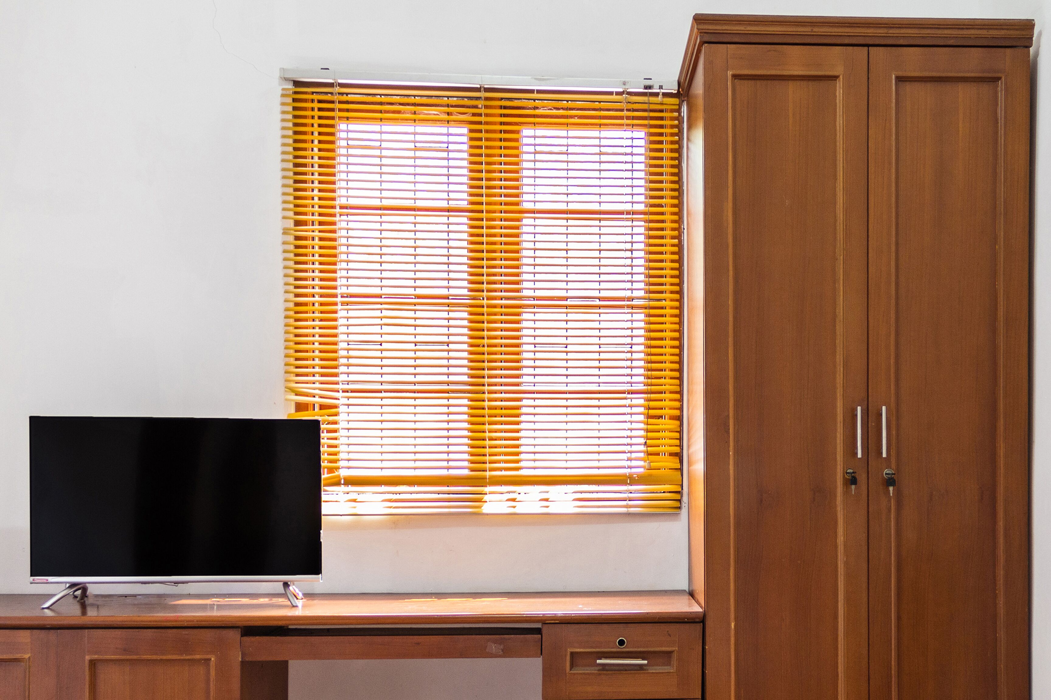 A room with a TV on a wooden desk, a window with yellow blinds, and a wooden wardrobe.