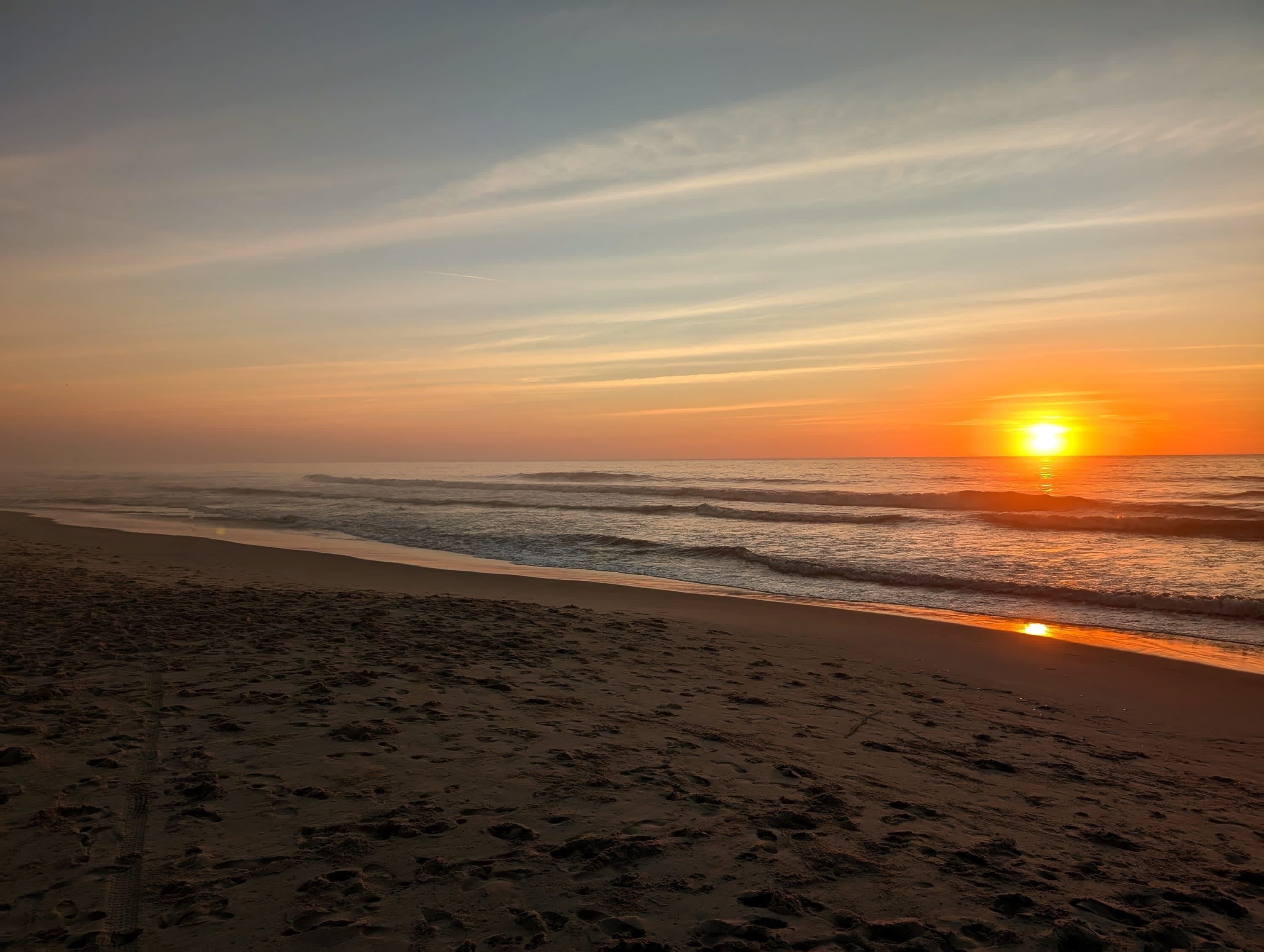 A beach with footprints leading to the water, a sunset in the background, and a clear sky.