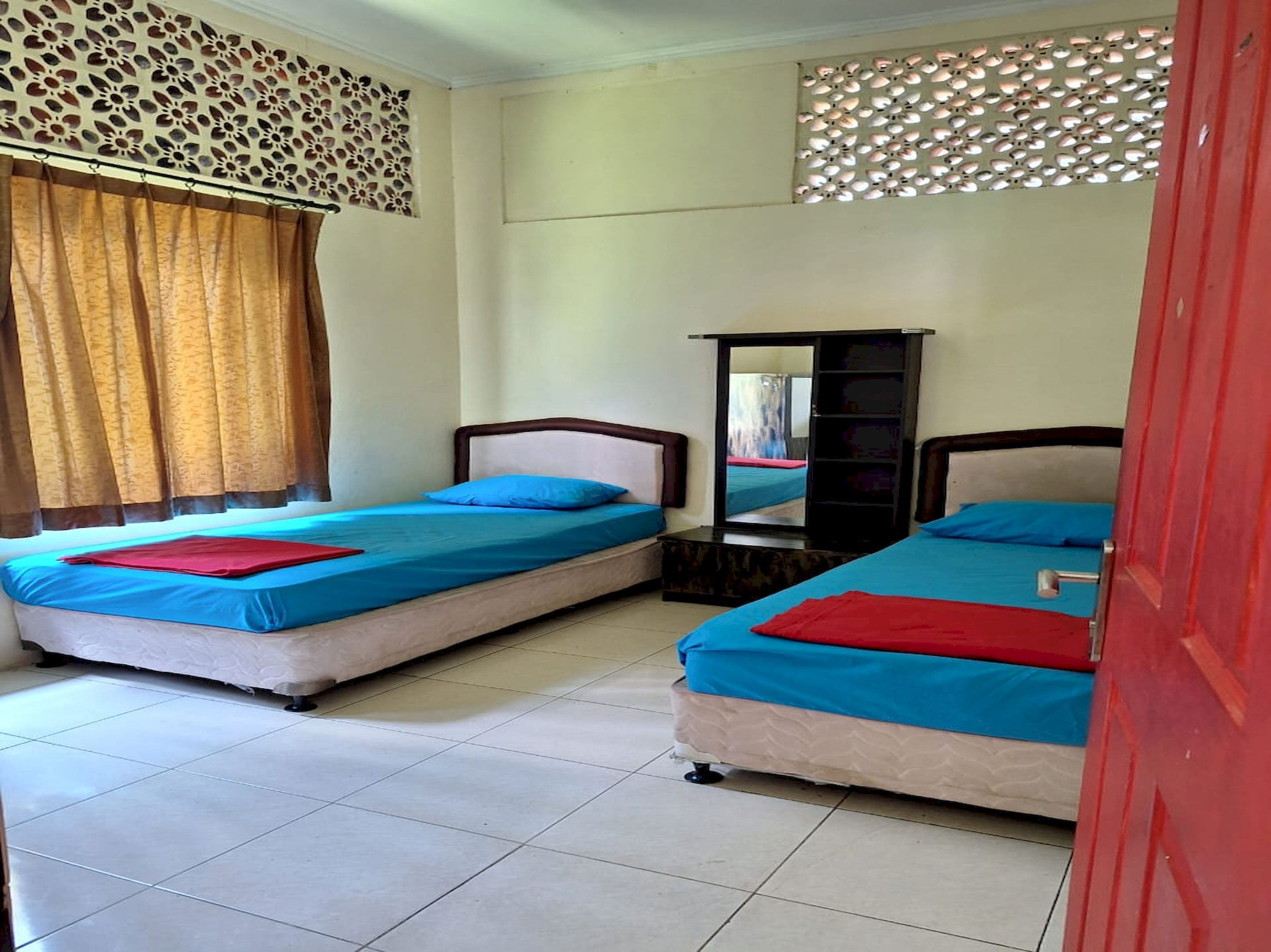 A room with two beds, each with blue bedding and red pillows. There is a mirror on the wall and a window with curtains.