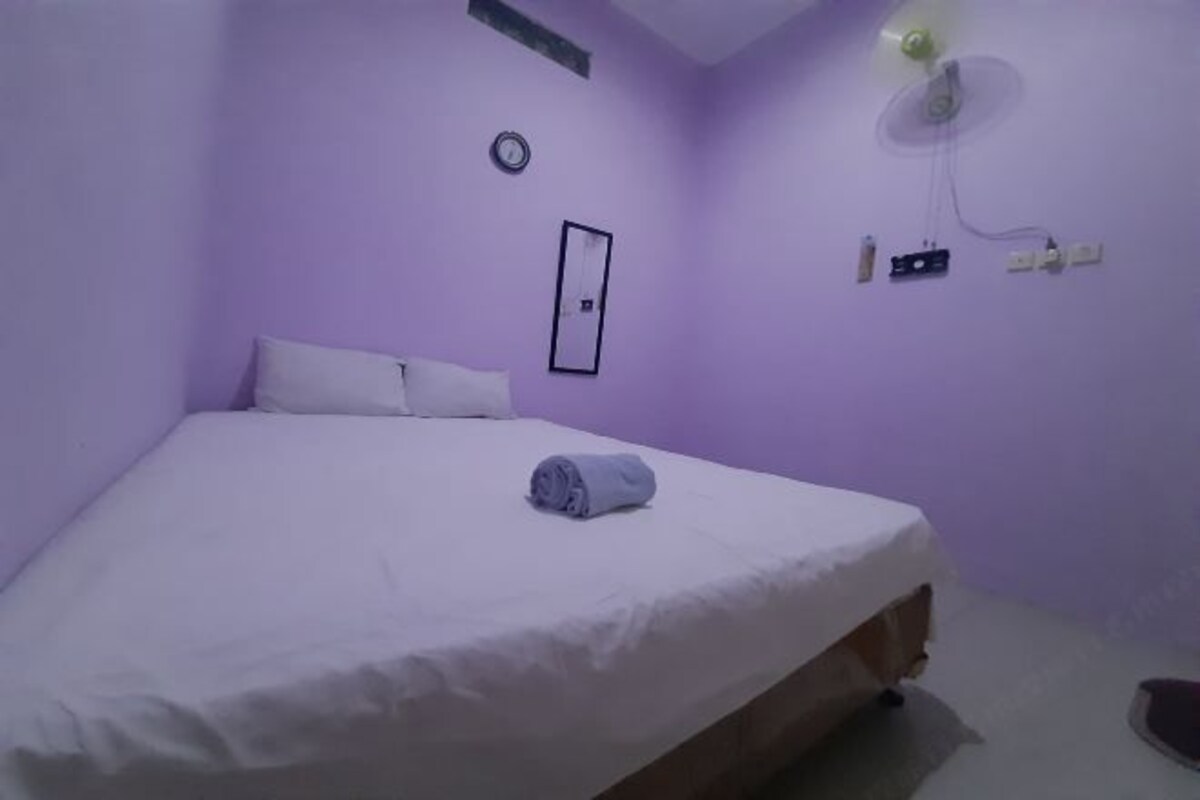 A single-bed room with a white bedspread, a folded towel, a wall clock, a framed picture, and a ceiling fan.