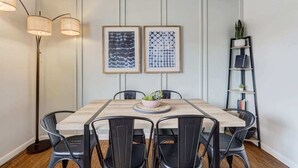 Een eethoek met een houten tafel, zwarte stoelen, twee ingelijste kunstwerken, een vloerlamp en een boekenkast met een plant.