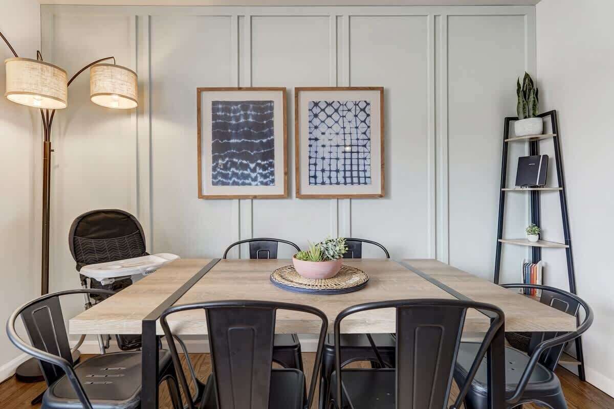 Un coin repas avec une table en bois, des chaises noires, deux œuvres d’art encadrées, une plante en pot et une bibliothèque.