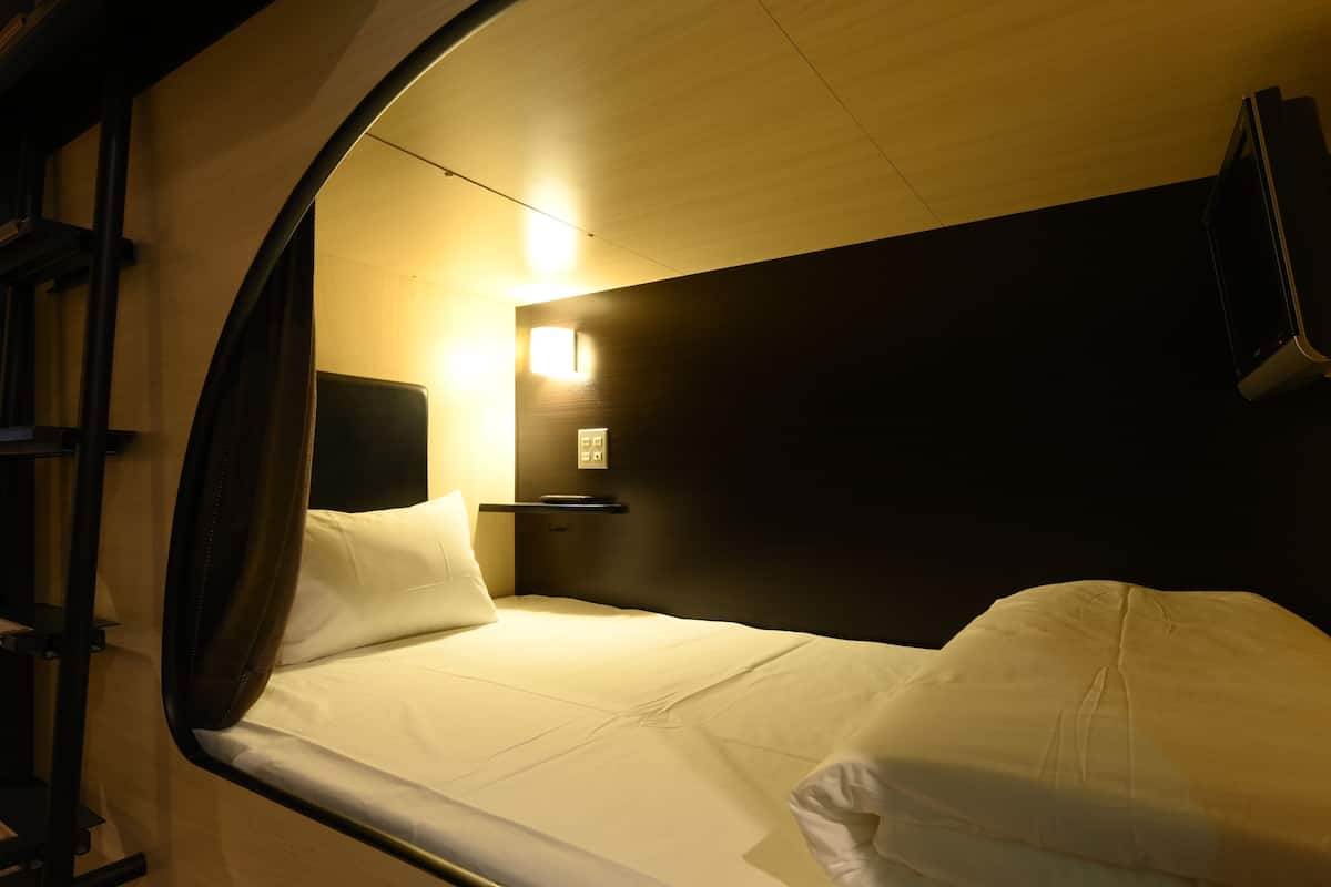 A compact, single-bed room with a curved wall, a bedside table, a lamp, and a wall-mounted TV.