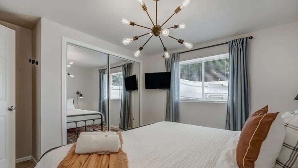 Une chambre à coucher avec un grand lit, un lustre moderne, une télévision et une salle de bain visible à travers une porte ouverte.