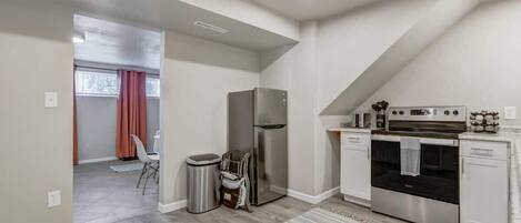 A modern kitchen with a refrigerator, oven, and microwave. There is a trash can and a small table with chairs. The room has a sloped ceiling and a window with curtains.