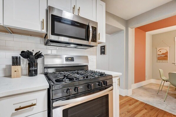 A modern kitchen with white cabinets, a stainless steel microwave, a gas stove, and a built-in oven. There is a knife block on the countertop and a chair in the adjacent dining area.