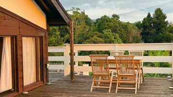 A wooden deck with a table and chairs, a white fence, and a building with a brown roof and windows.