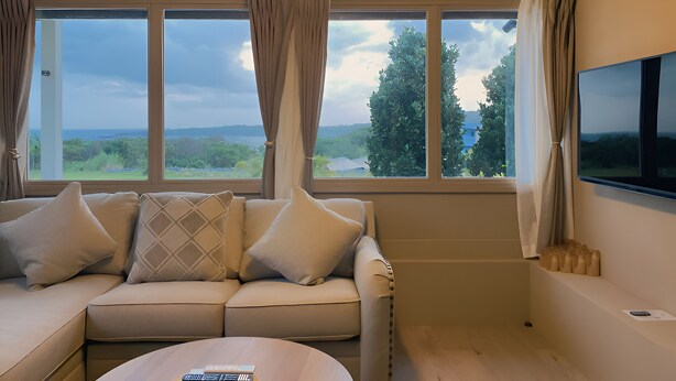 A modern living room with a beige sofa, a flat-screen TV, and large windows offering a view of the outdoors. - Villa SERENO MOTOBU  (Motobu)