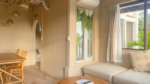 A cozy living room with a wooden dining table, a round coffee table with a remote control and papers, a beige sofa, and a large window with a view of greenery outside. - Villa SERENO MOTOBU  (Motobu)