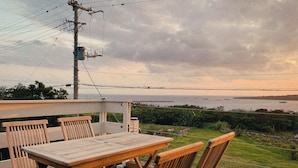 A wooden deck with a table and chairs, overlooking a body of water with boats and a distant shoreline. - Villa SERENO MOTOBU  (Motobu)