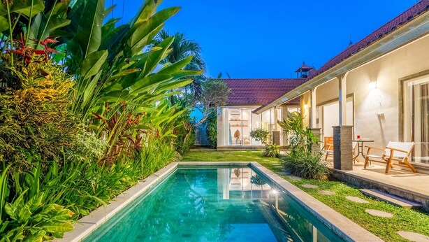 En swimmingpool omgivet af frodigt grønt, en overdækket terrasse med siddepladser, og et hus med et tag af fliser.