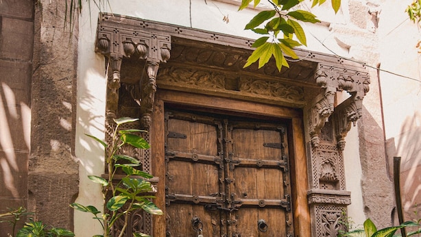 Une porte en bois rustique, ornée de sculptures complexes, flanquée de plantes en pot et d’un mur de pierres.